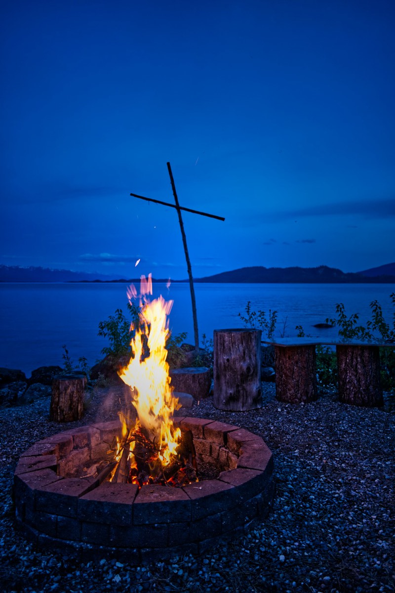 A fire pit burning at blue dusk beside calm coastal water with log seats (placeholder photo)