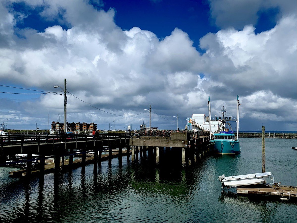 A harbour pier stacked with crab pots and a moored fishing boat under big clouds (placeholder photo)